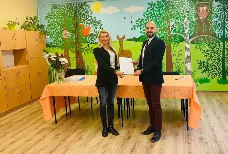 Zdjęcie wykonane w orientacji poziomej przedstawia kobietę i mężczyznę stojących w sali przedszkolnej przed stołem przykrytym pomarańczowym obrusem. Kobieta po lewej stronie ma na sobie czarną marynarkę, dżinsy i ciemne botki, natomiast mężczyzna po prawej jest ubrany w ciemną marynarkę, białą koszulę, ciemny krawat i bordowe spodnie. Wspólnie trzymają biały dokument, który jest głównym punktem centralnym tej fotografii. Tło stanowi kolorowy mural przedstawiający leśny krajobraz z drzewami, sarną, sową i ptakami, co nadaje wnętrzu edukacyjny charakter. Po lewej stronie kadru widoczne są wysokie, jasne szafy meblowe oraz wazon z bukietem czerwonych i białych kwiatów ustawiony na stole. Cała kompozycja jest symetryczna i dobrze oświetlona, co czyni ją odpowiednią do publikacji dotyczących współpracy społecznej lub edukacyjnej.