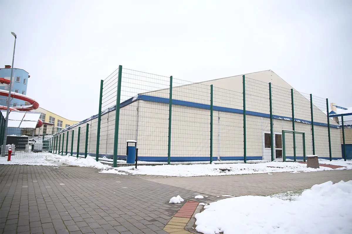 Zdjęcie w orientacji poziomej przedstawia zadaszoną halę sportową o konstrukcji namiotowej, otoczoną wysokim zielonym ogrodzeniem z siatki. Obiekt znajduje się na otwartym terenie, a podłoże wokół niego pokryte jest miejscami warstwą śniegu oraz szarą kostką brukową. Po lewej stronie widoczna jest część niebieskiego budynku z czerwoną, spiralną zjeżdżalnią, co sugeruje lokalizację w pobliżu parku wodnego lub centrum rekreacyjnego. Jasne ściany hali z niebieskimi akcentami wyraźnie kontrastują z szarym, zimowym niebem. Kompozycja zdjęcia obejmuje cały budynek hali, podkreślając jego surową, użytkową architekturę w otoczeniu zimowego krajobrazu.